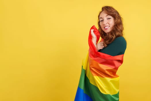 Happy redheaded woman wrapped with a lgbt flag in studio with yellow background