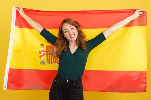 Happy redheaded woman smiling and raising a spanish national flag in studio with yellow background
