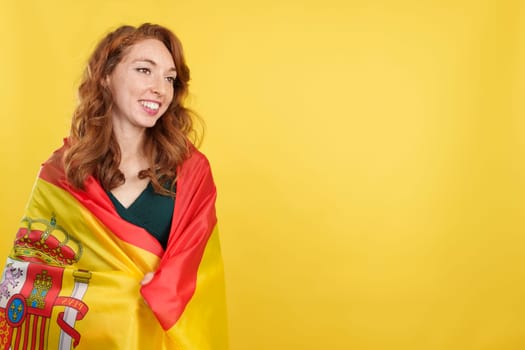 Happy redheaded woman wrapping with a spanish national flag in studio with yellow background