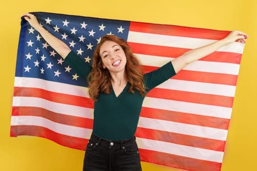 Happy redheaded woman raising a north america national flag in studio with yellow background