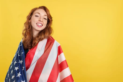 Happy redheaded woman wrapping with a north america national flag in studio with yellow background