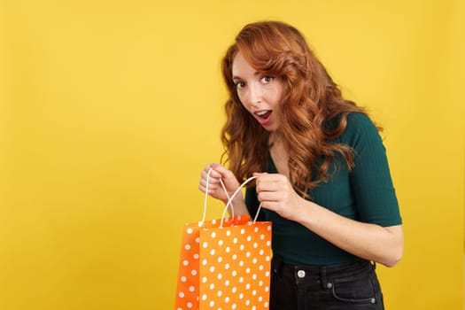 Surprised redheaded woman with a shopping bag in studio with yellow background