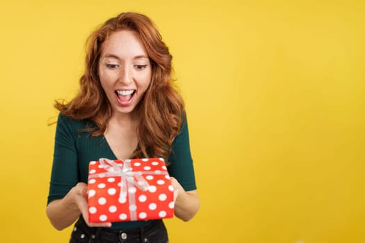 Excited redheaded beauty woman holding a gift in studio with yellow background