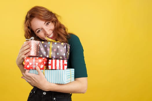 Redheaded woman embracing many gifts while smiling at camera in studio with yellow background
