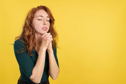 Redheaded woman praying with folded hands and eyes closed in studio with yellow background