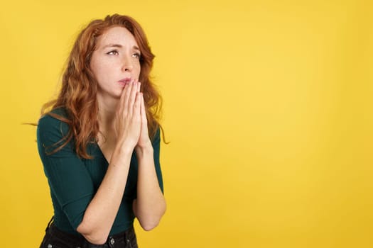 Redheaded woman looking up while praying with folded hands in studio with yellow background