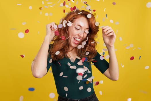 Happy redheaded woman dancing surrounded by confetti flying in the air in studio with yellow background