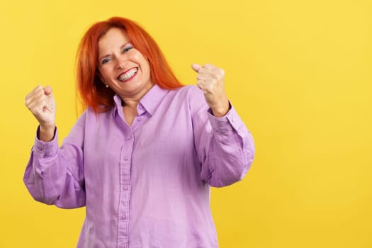 Happy mature woman with dye redhead celebrating while raising arms in studio with yellow background