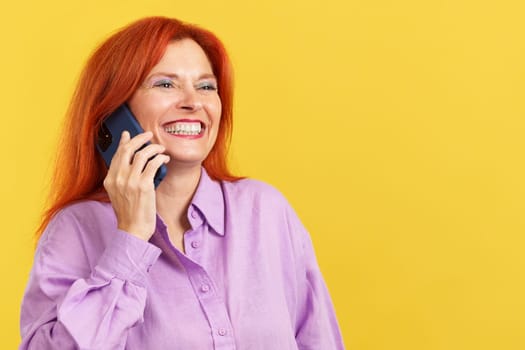 Happy mature woman with red dye hair talking to the mobile in studio with yellow background