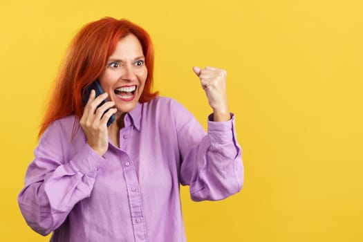 Happy mature redheaded woman celebrating while taking to the mobile in studio with yellow background