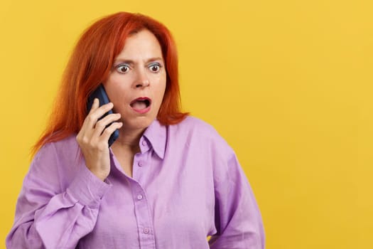 Surprised mature redheaded woman talking to the mobile in studio with yellow background