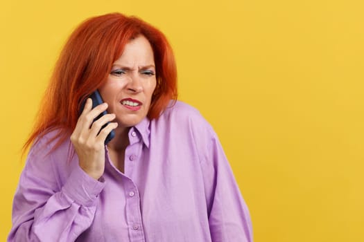 Angry redheaded mature woman talking to the mobile in studio with yellow background