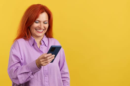 Happy mature redheaded woman smiling while using the mobile in studio with yellow background