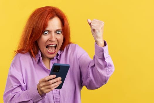 Excited mature redheaded woman celebrating while using the mobile phone in studio with yellow background