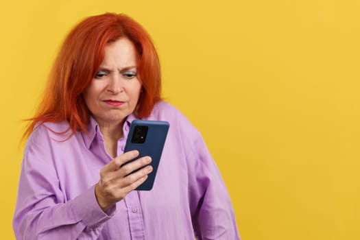 Worried redheaded mature woman using a mobile phone in studio with yellow background