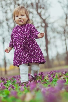 blond baby in a colored dress running with her tongue out in a forest glade with flowers