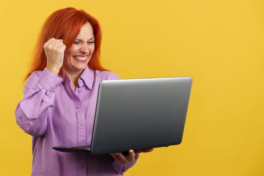 Mature redheaded woman celebrating while looking at the laptop in studio with yellow background