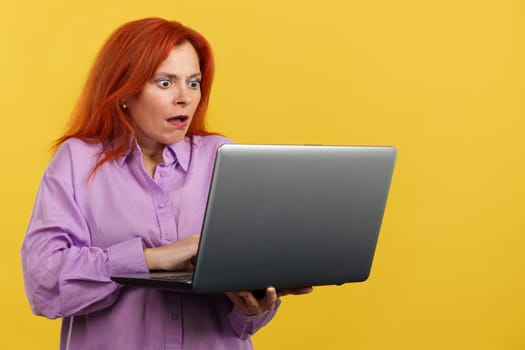 Redheaded mature woman with a surprised look using a laptop in studio with yellow background