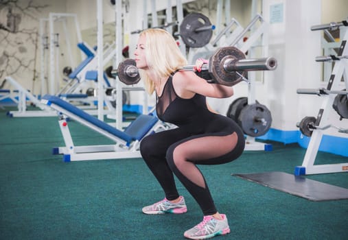 Slim girl in black tight jumpsuit squat with a barbell.