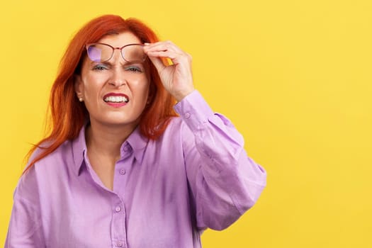 Aged redheaded woman taking off her glasses to see in studio with yellow background