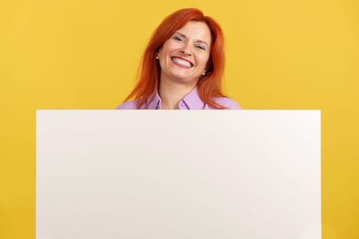 Mature redheaded woman holding a blank panel while smiling at the camera in studio with yellow background