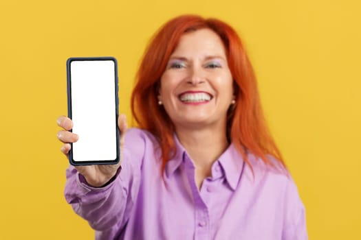 Happy redheaded mature woman holding a mobile with a blank screen in studio with yellow background