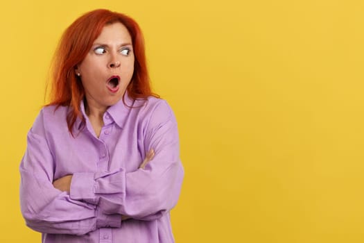 Surprised redheaded mature woman looking away with the mouth opened in studio with yellow background