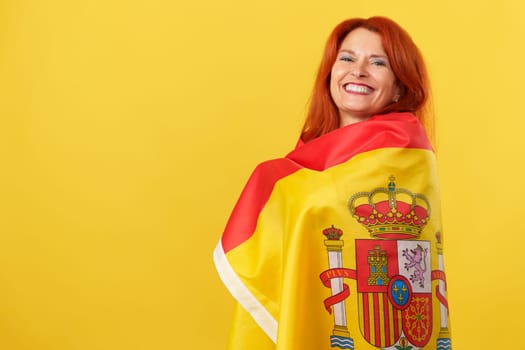 Happy redheaded mature woman wrapping with a spanish national flag in studio with yellow background