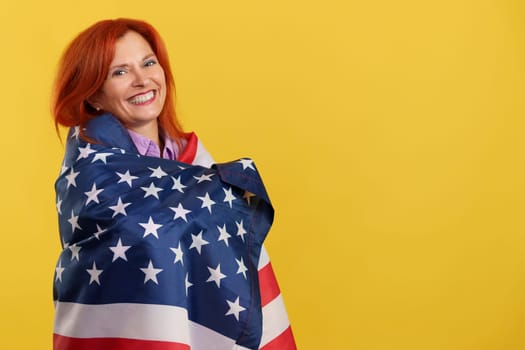 Happy redheaded mature woman wrapping with a north america national flag in studio with yellow background