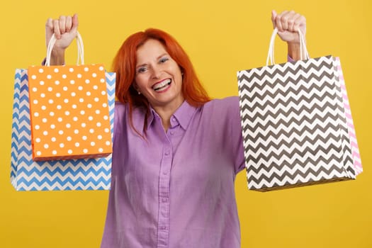 Mature redheaded woman raising many shopping bags while smiling at camera in studio with yellow background