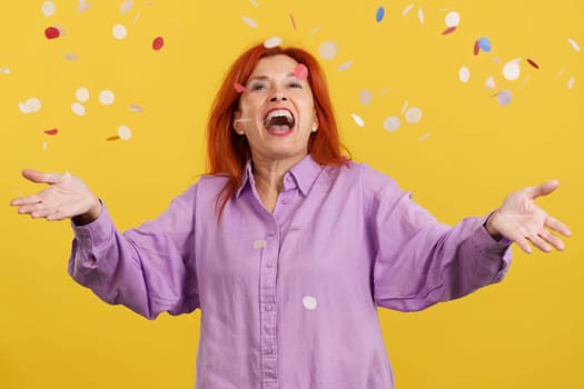 Happy redheaded mature woman surrounded by confetti flying in the air in studio with yellow background