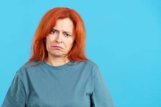 Mature redheaded woman looking at camera with sad expression in studio with blue background