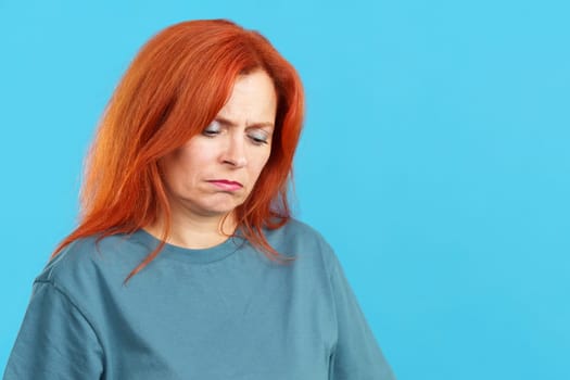 Mature redheaded woman looking down with sad expression in studio with blue background