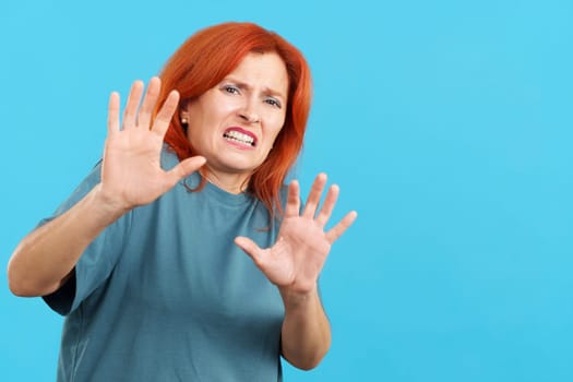 Redheaded mature woman gesturing with the hands in fear in studio with blue background