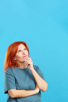 Redheaded mature woman with hand on face and thoughtful expression in studio with blue background