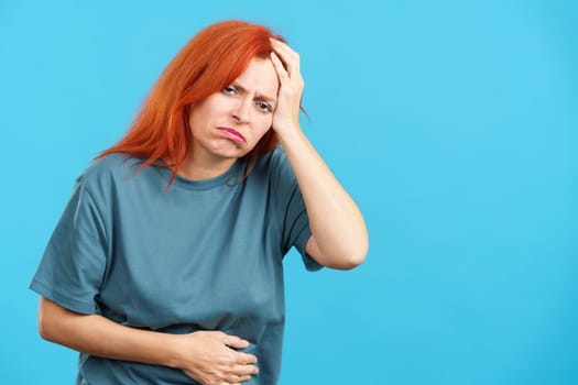 Mature redheaded woman gesturing bodily discomfort and headache in studio with blue background