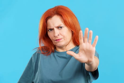 Redheaded mature woman gesturing prohibition with hand in studio with blue background