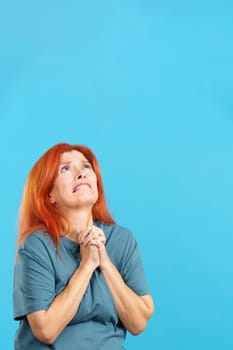 Redheaded mature woman praying in despair looking up with folded hands in studio with blue background