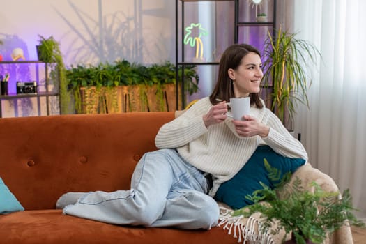 Portrait of attractive smiling young woman drinking a cup of warm coffee or herbal tea at home living room apartment in the morning. Pretty girl lying on sofa enjoying comfortable relaxing alone