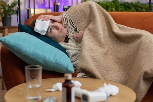 Young ill woman suffering from cold or allergy sitting on sofa in living room at home. Sick girl wear hat wrapped in plaid blows sneezes wipes snot into napkin. Coronavirus quarantine, pandemic