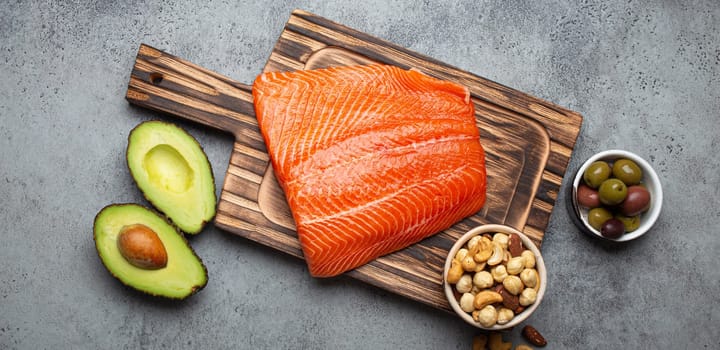 Food sources of healthy unsaturated fat: fresh raw salmon fillet, avocado, olives, nuts on cutting board on rustic stone background top view. Omega 3 and oil products, healthy nutrition and keto diet