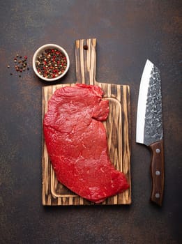 Raw uncooked top round beef steak on wooden cutting board with big kitchen knife and pepper on dark brown rustic stone background top view, cooking meat steak concept