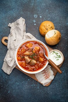 Ukrainian Borscht, red beetroot soup with meat, in white bowl with sour cream, garlic buns Pampushka and salo slices on rustic stone background. Traditional authentic dish of Ukraine