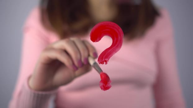 A young woman draws a question mark on the glass. A girl in a pink sweater draws with a brush and red paint. Close-up. 4k