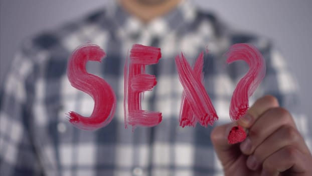 A young man draws on the glass the word sex with a question mark. A man in a plaid shirt draws with a brush and red paint. Close-up. 4k