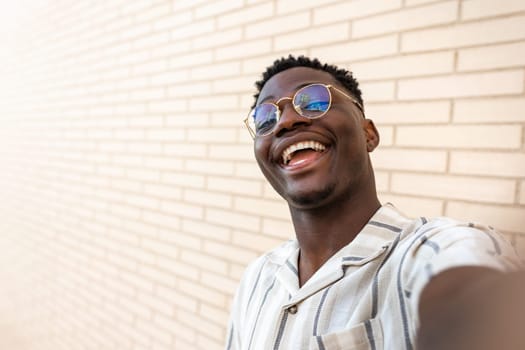 Happy, smiling young African American man with glasses taking selfie. Copy space. Brick wall background. Lifestyle concept.