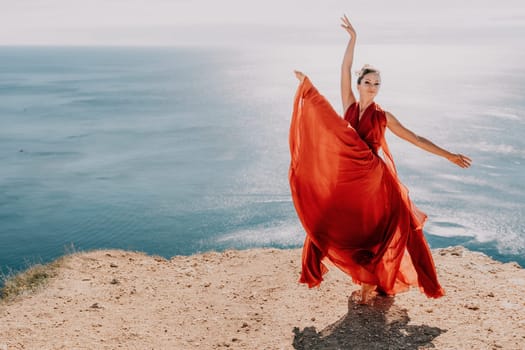 Side view a Young beautiful sensual woman in a red long dress posing on a rock high above the sea during sunrise. Girl on the nature on blue sky background. Fashion photo.