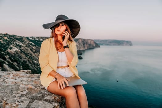 Successful business woman in yellow hat working on laptop by the sea. Pretty lady typing on computer at summer day outdoors. Freelance, travel and holidays concept.