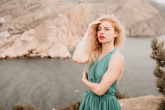 Side view a Young beautiful sensual woman in a mint long dress posing on a volcanic rock high above the sea during sunset. Girl on the nature on overcast sky background. Fashion photo