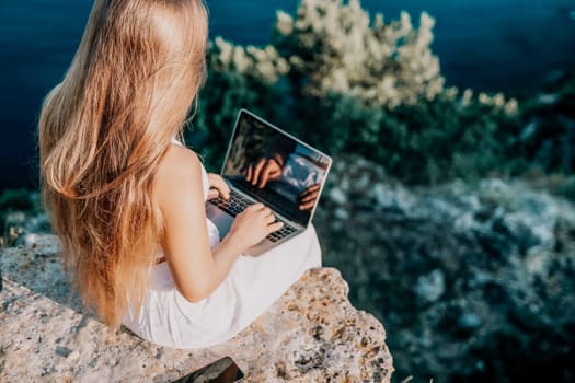 Successful business woman in yellow hat working on laptop by the sea. Pretty lady typing on computer at summer day outdoors. Freelance, travel and holidays concept.